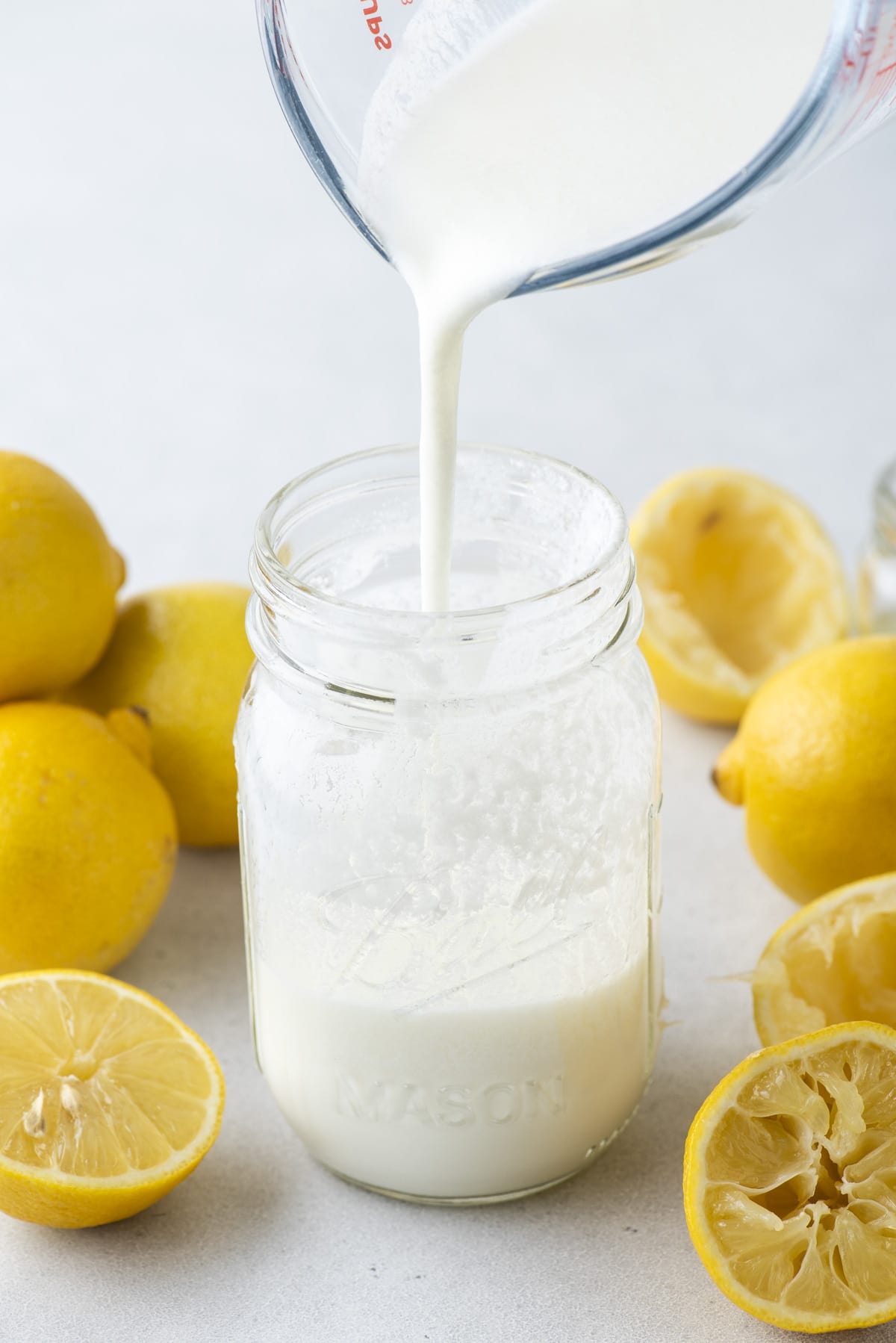a mason jar with buttermilk from a clear class measuring cup being poured into the jar and fresh whole and sliced lemons surrounding the jar