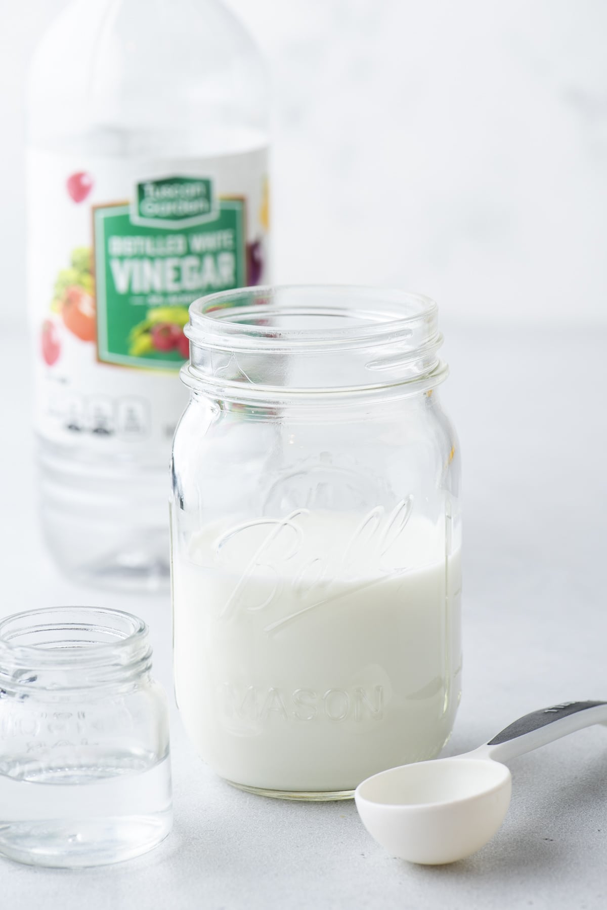 a clear class jar of buttermilk with a white tablespoon and a small clear glass jar of vinegar and a bottle of vinegar in the background
