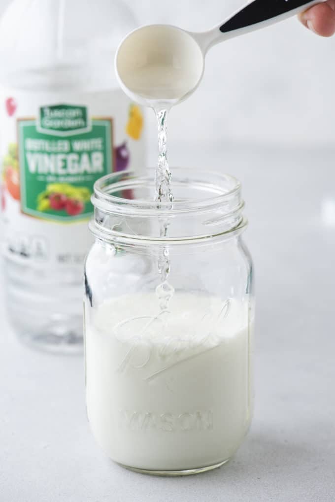 a mason jar with buttermilk in it with a tablespoon of vinegar being poured in and a bottle of vinegar behind it