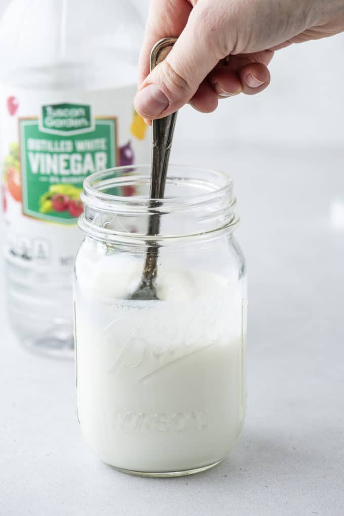 a clear class jar of buttermilk being stirred with a spoon, with a bottle of white vinegar in the background