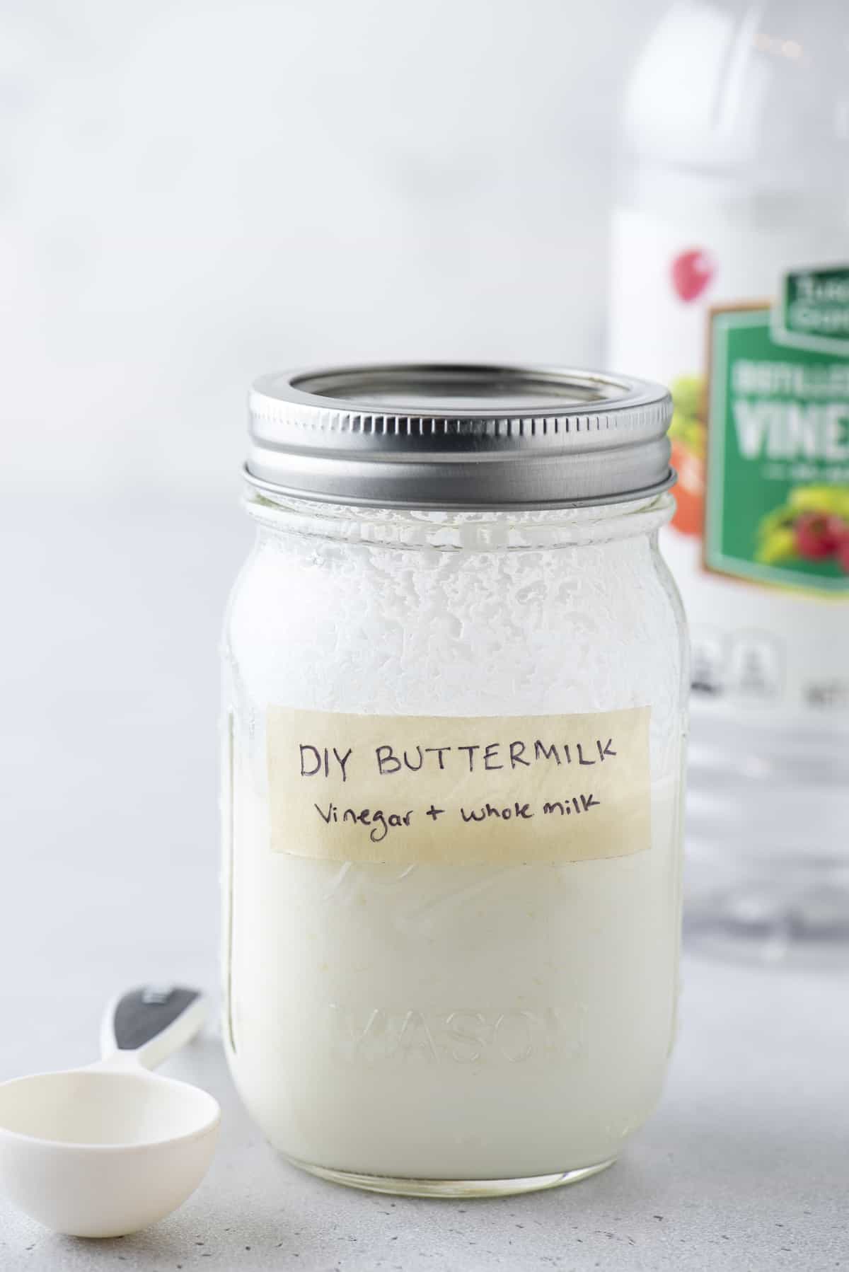 a mason jar full of buttermilk with a lid on, labeled with tape that says "DIY Buttermilk: vinegar + whole milk", with a measuring spoon beside it an a bottle of vinegar in the background