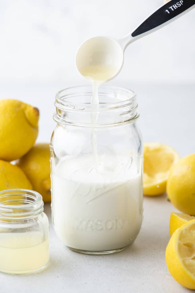 a mason jar of buttermilk with a tablespoon of lemon juice being poured in, a small clear glass jar of lemon juice beside it, surrounded by fresh whole and sliced lemons