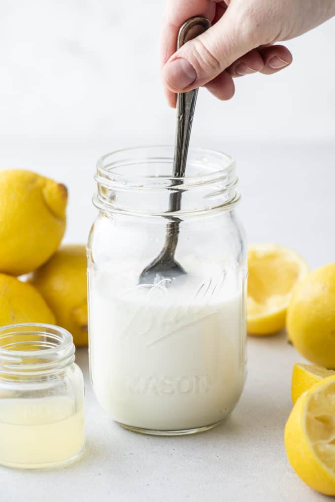 a mason jar with buttermilk in it being stirred, a small clear glass jar of lemon juice beside it and whole and sliced lemons surrounding it