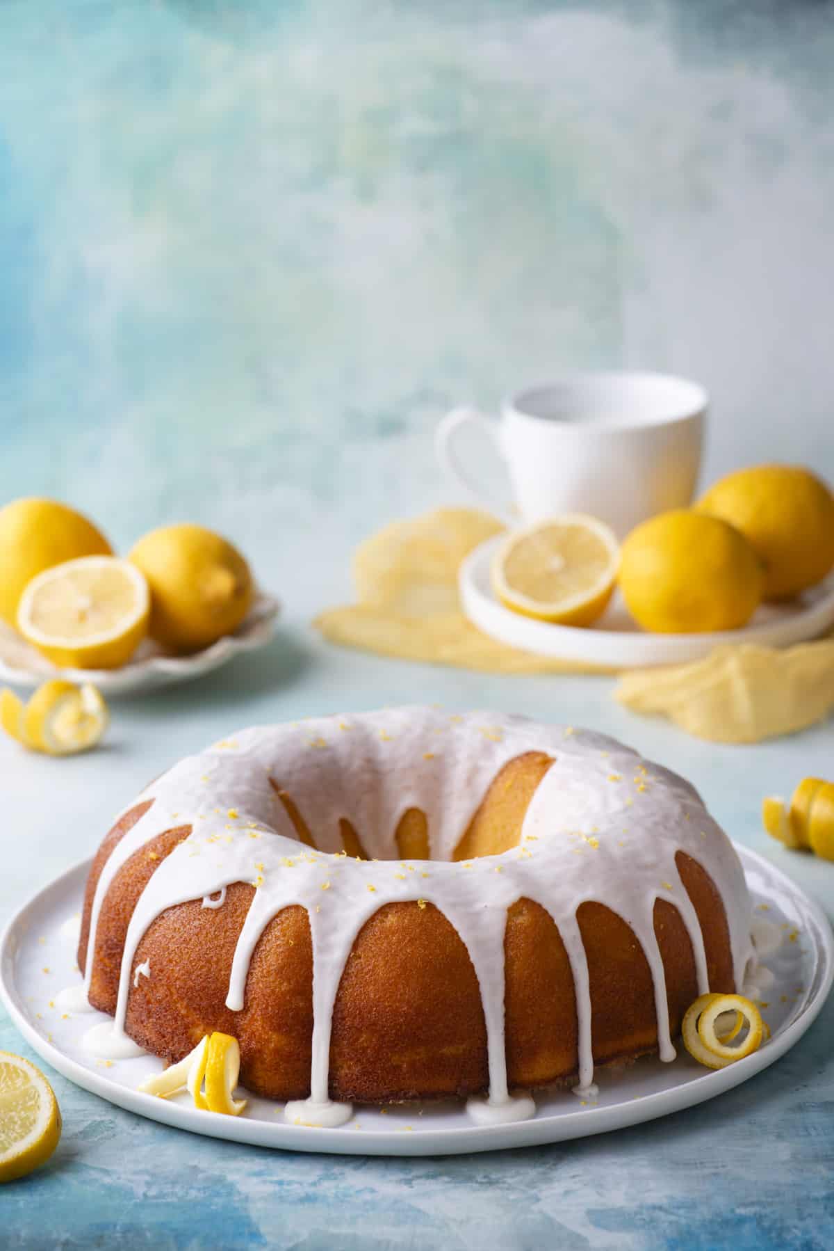 a lemon bundt cake on a round, white plate with fresh lemon peels around it, sitting on a blue surface with small white places of fresh lemons and more lemon peels and slices around it