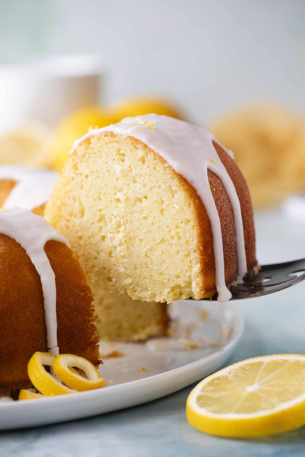a slice of lemon bundt cake being lifted away from the rest of the cake off a white plate sitting on a blue surface with a fresh lemon slice beside it