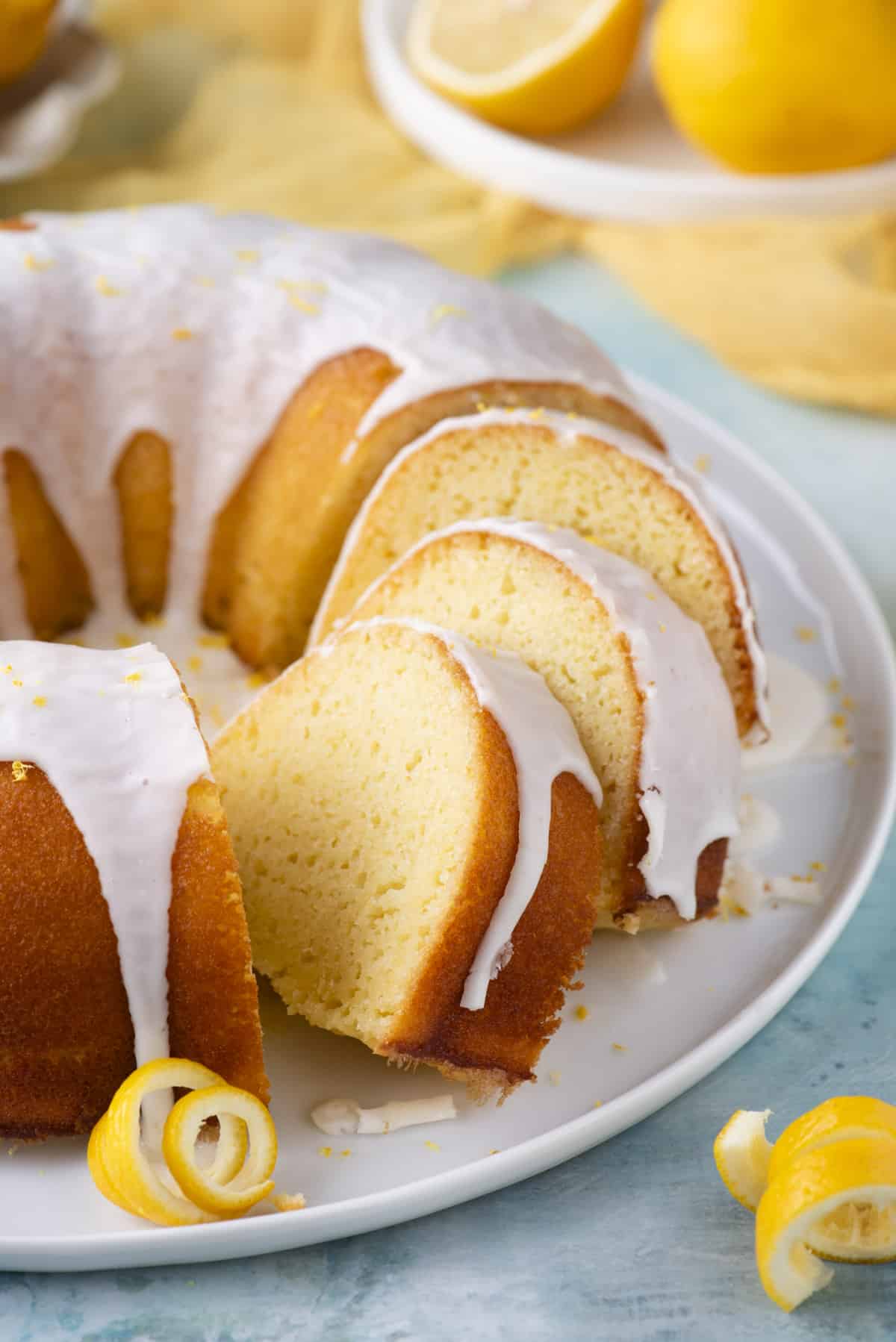 a lemon bundt cake on a round, white plate with three slices cut and the rest of the cake whole, surrounded by fresh lemon peels and a small white plate with fresh lemons on it