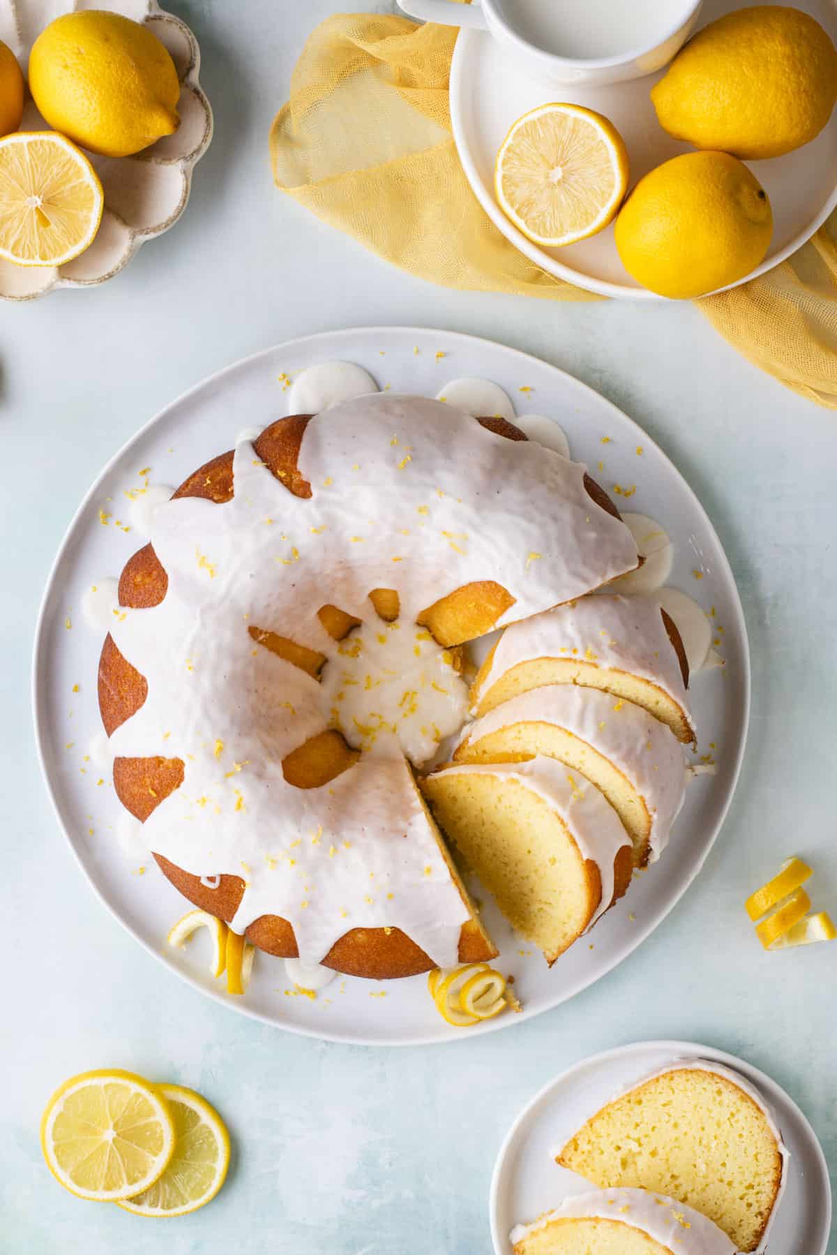 Over head view of a lemon bundt cake on a round, white plate with three pieces sliced and the rest of the cake whole, surrounded by other small white plates, one with two slices of lemon bundt cake, and two with fresh lemons
