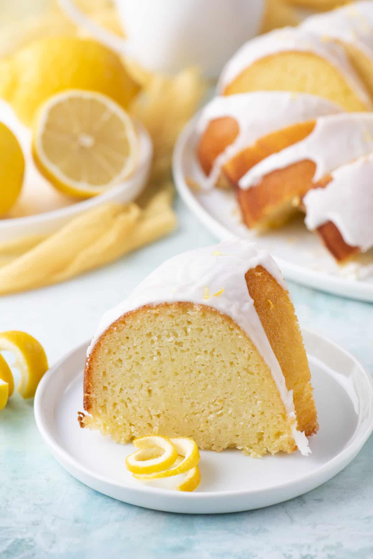 a slice of lemon bundt cake on a small, round white plate with a fresh lemon peel beside it and two plates in the background with more cake and fresh lemons on it