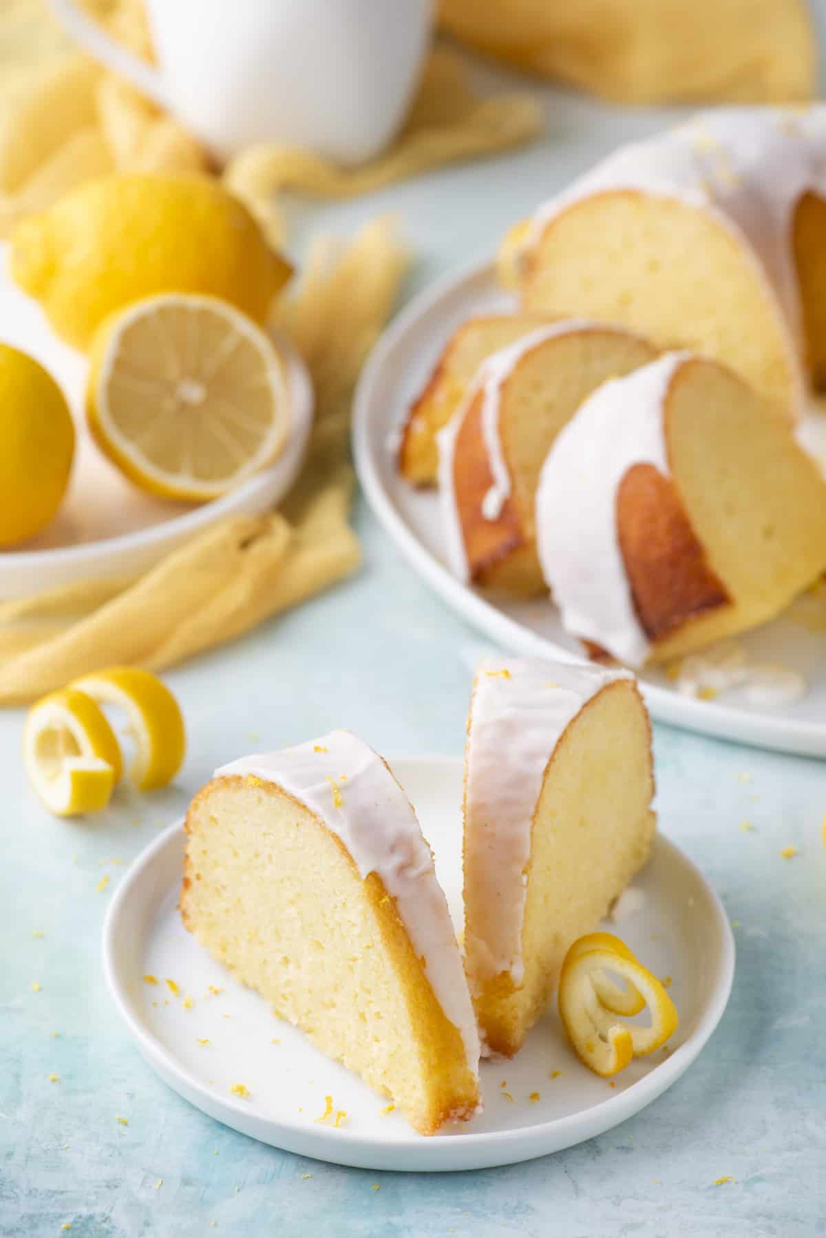 two slices of lemon bundt cake on a small white plate, surrounded by fresh lemon peels, a white plate with more slices of cake and another white plate with fresh lemons