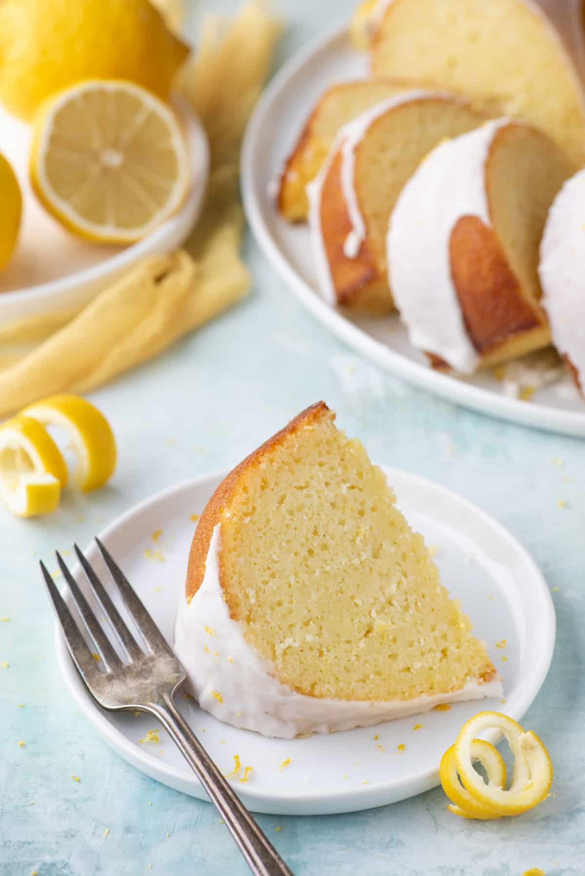 a slice of lemon bundt cake on a small white plate with a fork laying beside it, surrounded by fresh lemon peels, a white plate with more slices of cake and another white plate with fresh lemons