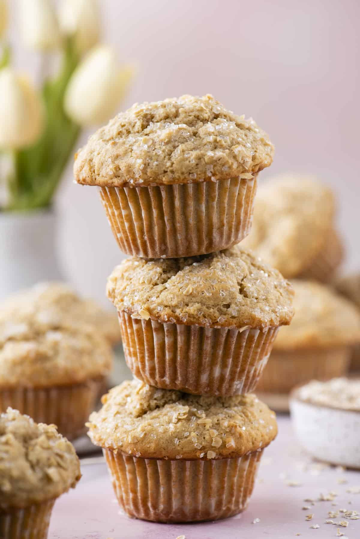 a stack of three oatmeal muffins on a light pink surface surrounded by more muffins and oats sprinkled around