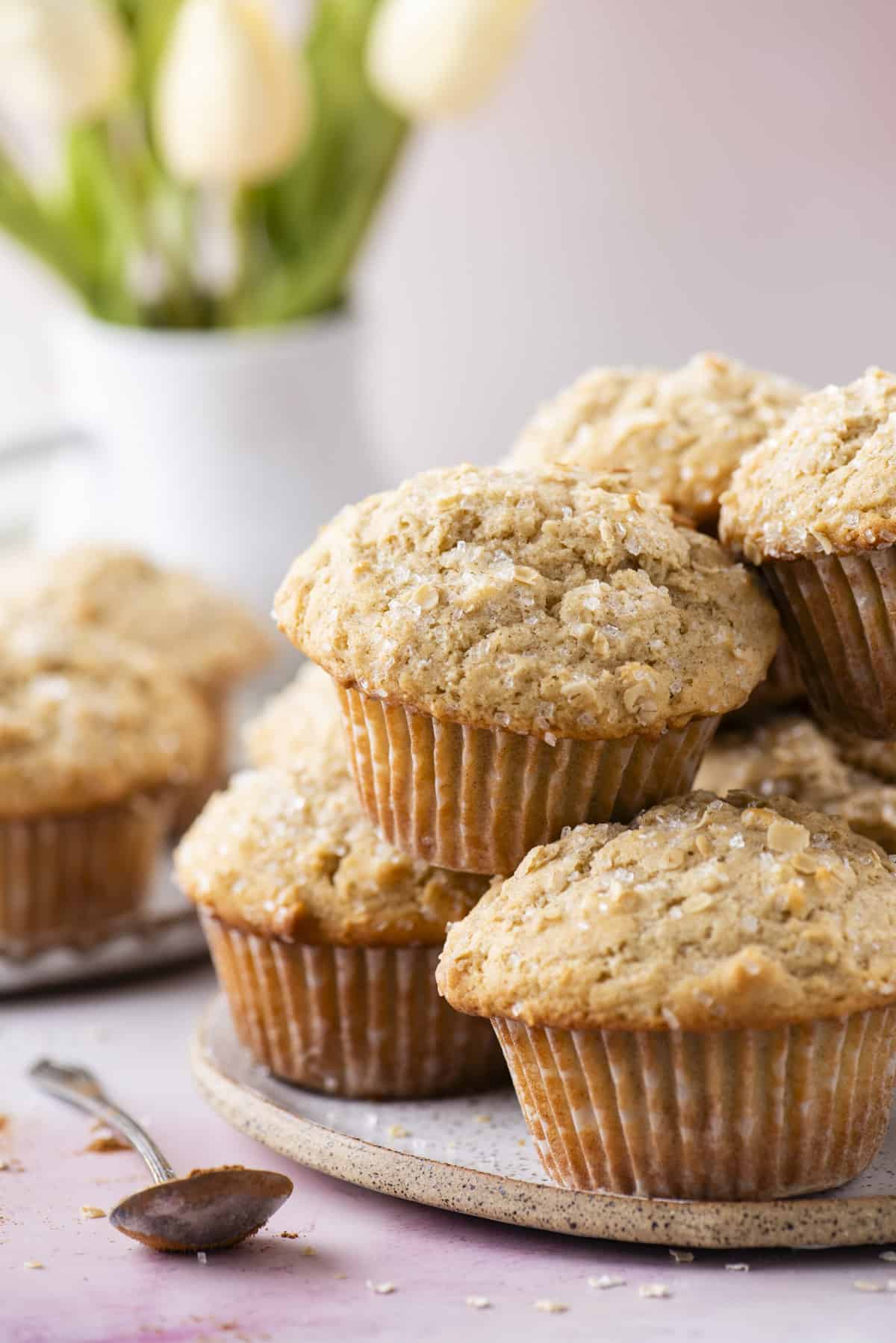 oatmeal muffins piled on top of a light grey speckled plate on a light pink surface with a spoon beside it and oats sprinkled around