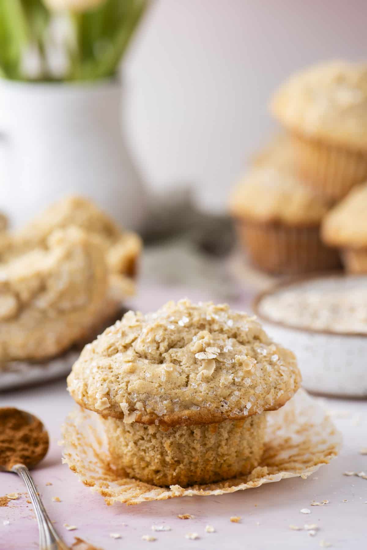 an oatmeal muffin sitting on top of its muffin liner that has been peeled away from the sides, with a spoon of cinnamon beside it, more muffins in the background and oats sprinkled around