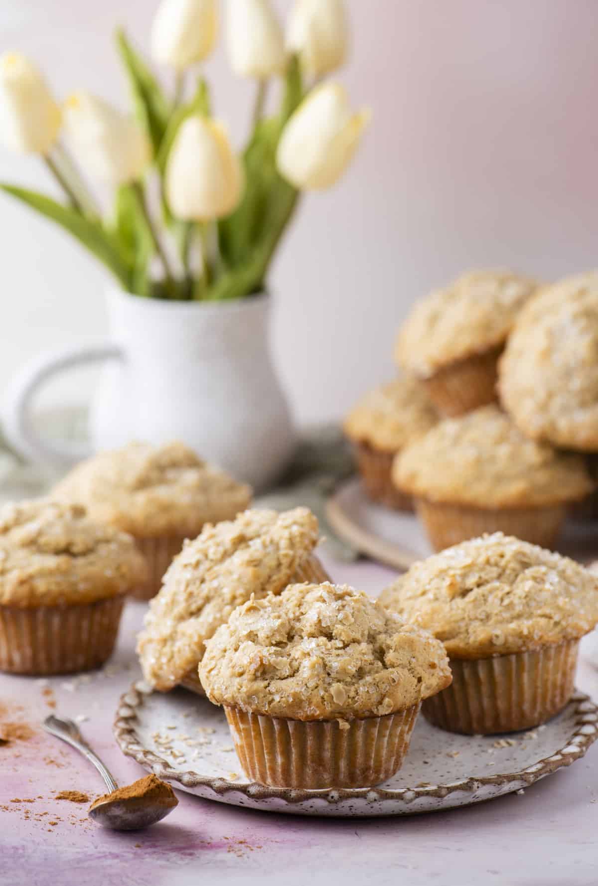 three oatmeal muffins on a light grey plate with a spoon of cinnamon beside it and more muffins and a vase of tulips in the background