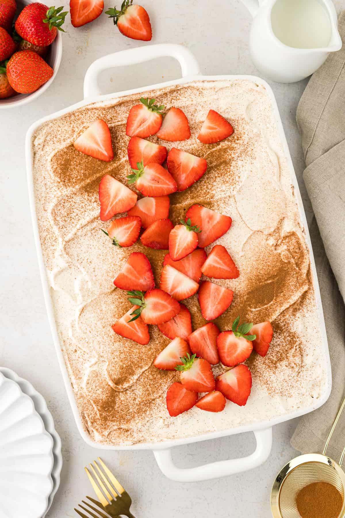 overhead view of a tres leches cake topping with cinnamon and fresh strawberries, surrounded by a sifter with cinnamon in it, two forks, two stacked white plates, a bowl of fresh strawberries and a cup of milk