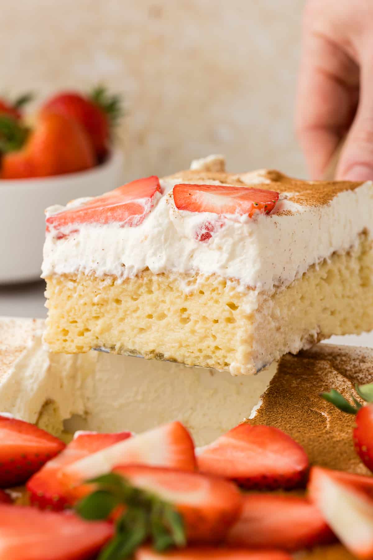 a slice of tres leches cake topped with fresh strawberries being lifted out of the dish away from the rest of the cake by a spatula