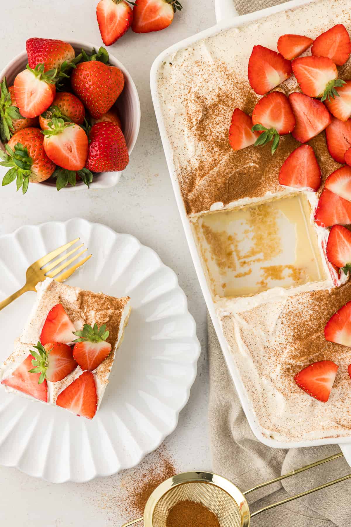 overhead view of a tres leches cake topped with fresh strawberries in a white baking dish with one slice missing, that slice beside it on a white plate, a sifter with cinnamon in it beside the plate and a bowl of fresh strawberries