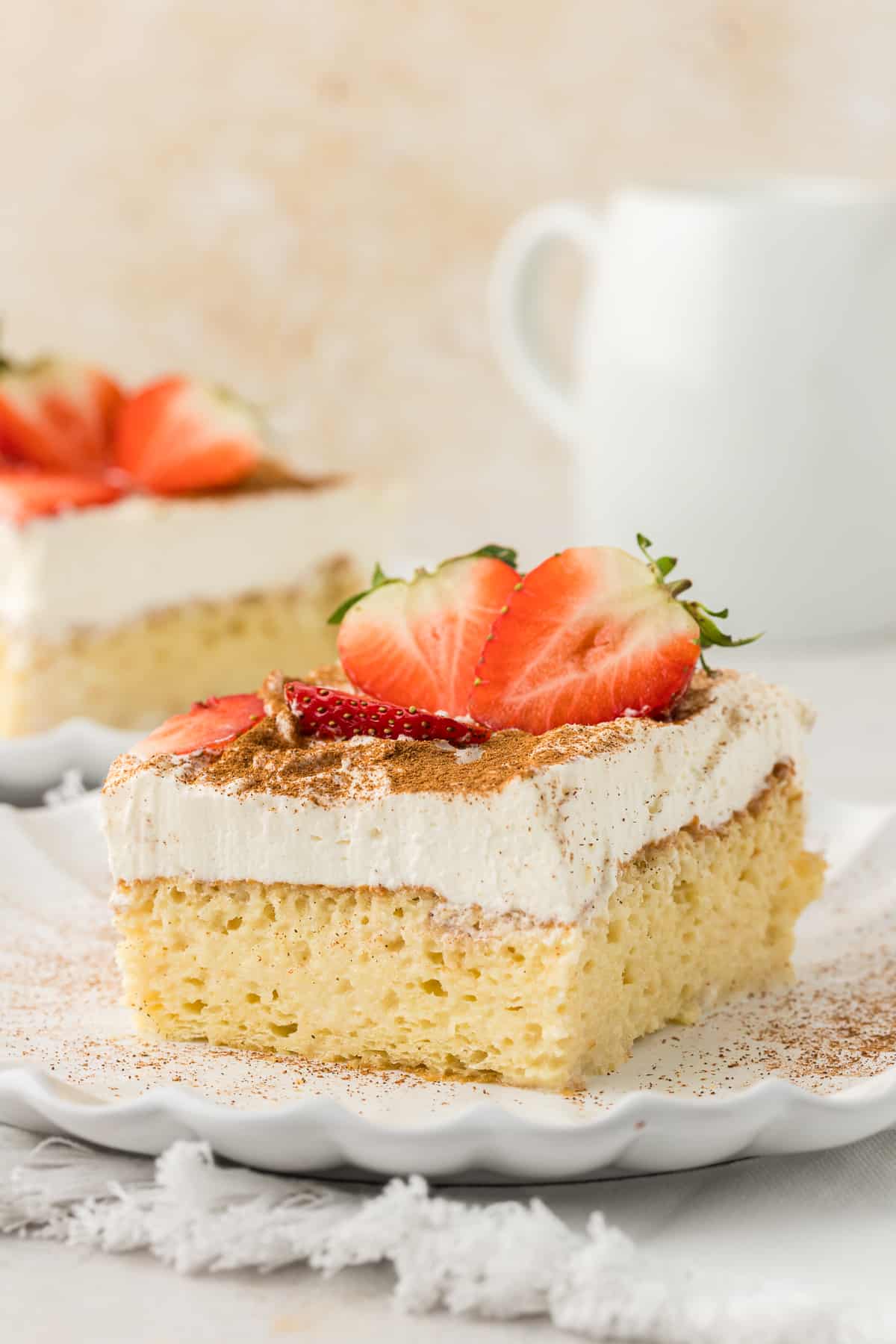 a slice of tres leches cake on a small white plate topped with fresh strawberry slices and another slice of cake on a plate in the background