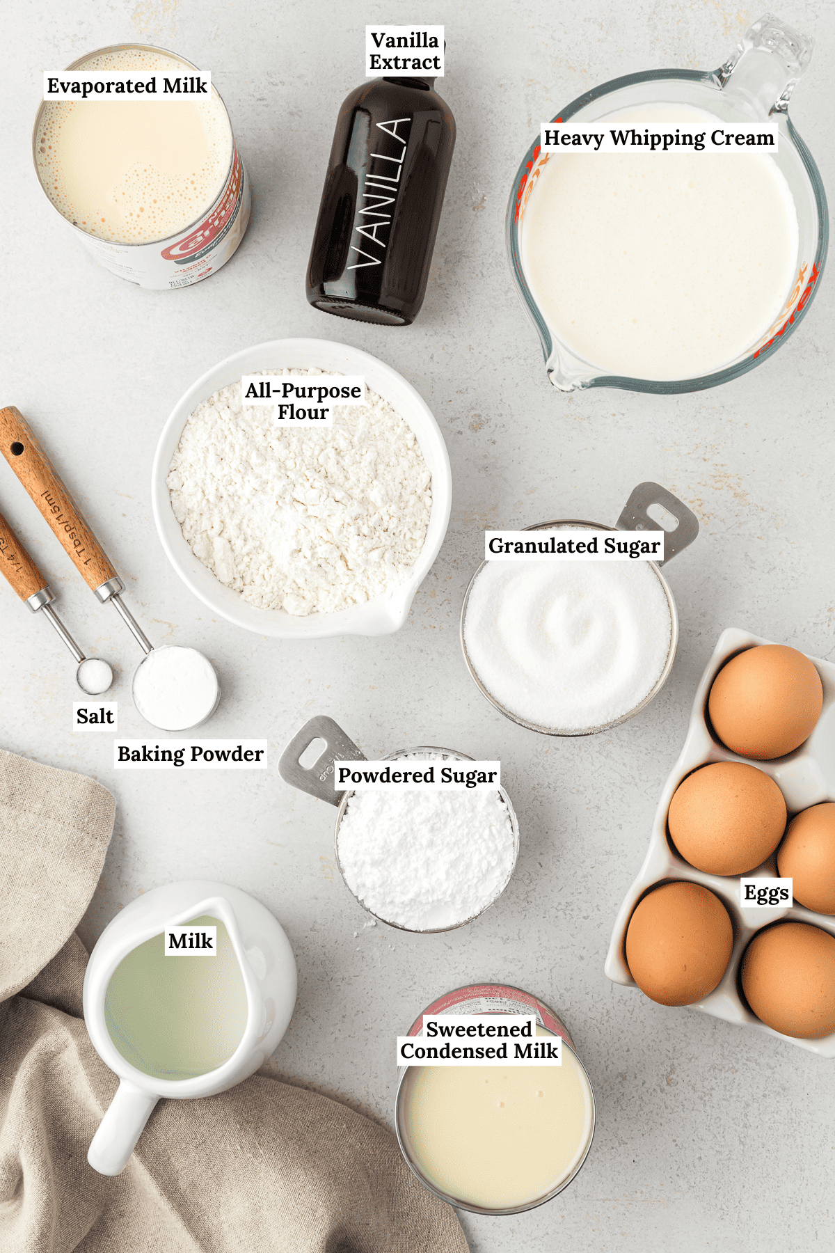 overhead view of the ingredients for tres leches cake including evaporated milk, vanilla extract, heavy whipping cream, all-purpose flour, salt, baking powder, granulated sugar, powdered sugar, milk, sweetended condensed milk, and eggs