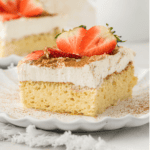 a slice of tres leches cake on a small white plate topped with fresh strawberry slices and another slice of cake on a plate in the background