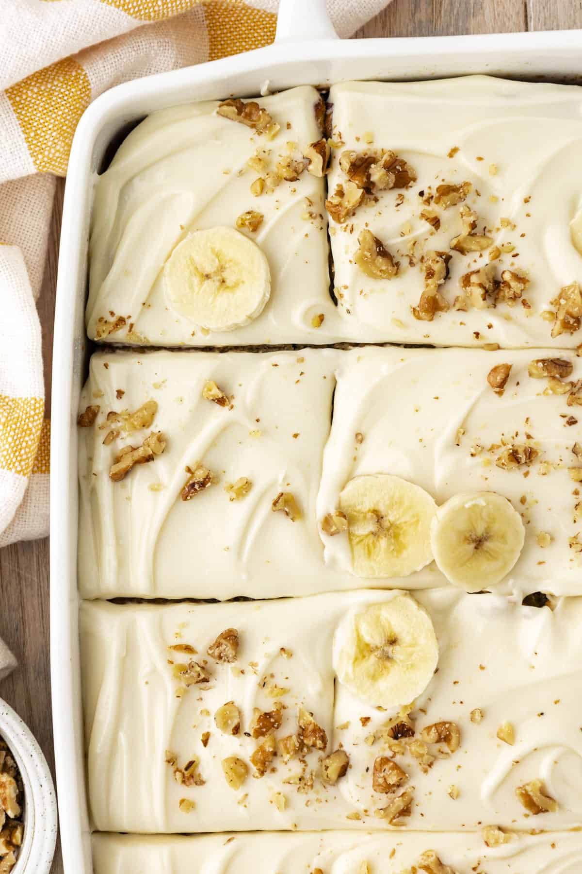 over head view of a banana cake in a white baking dish, sliced into pieces and topped with sliced bananas and chopped walnuts, with a yellow and white striped kitchen towel beside the pan