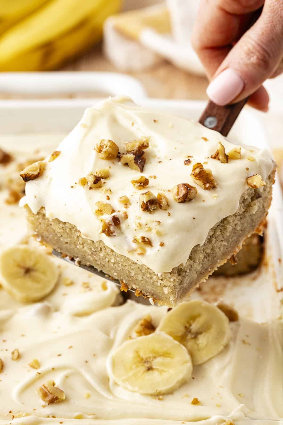 a slice of banana cake with chopped walnuts sprinkled on top of it being lifted out of the cake dish with a spatula
