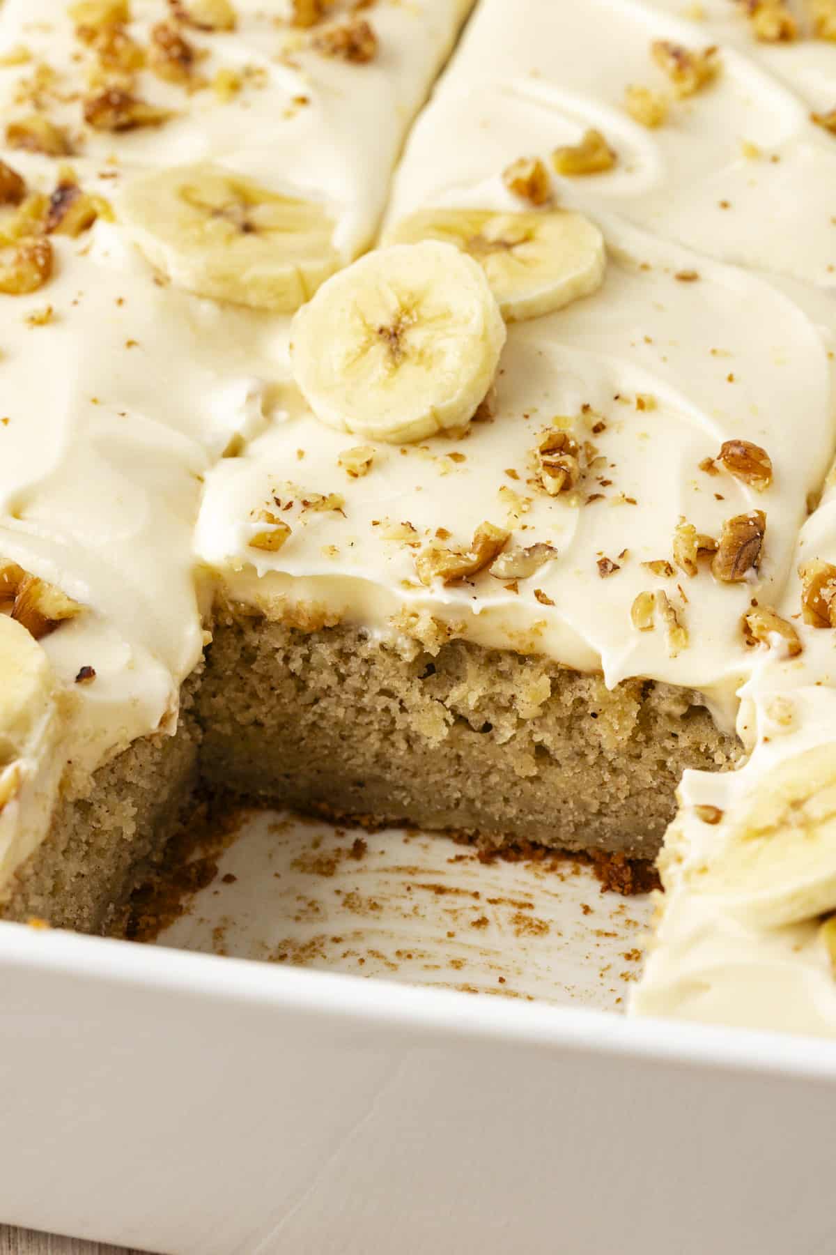 close up of a white baking dish full of banana cake with cream cheese frosting with one slice missing showing the inside with the layer of cake and layer of frosting on top