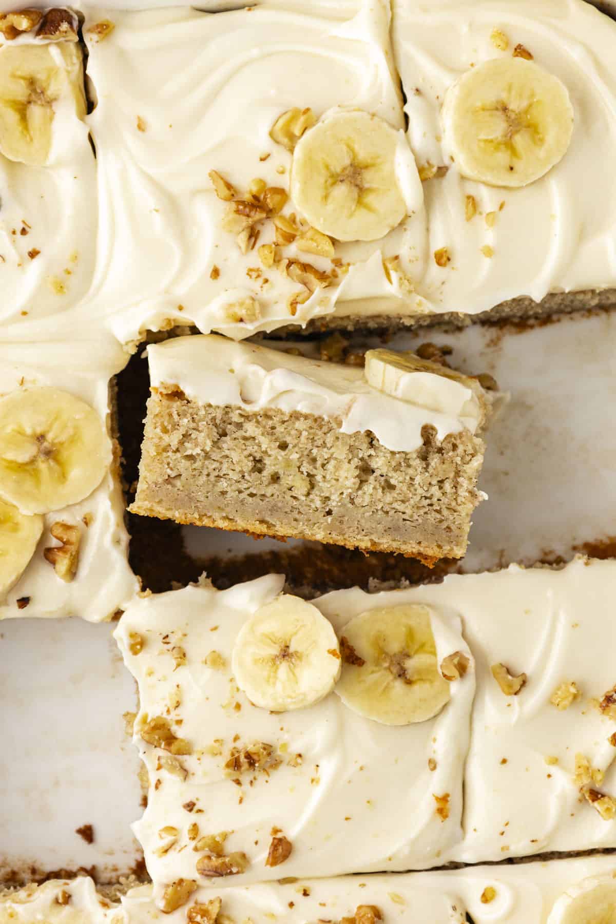 close up of a banana cake with some slices missing and one slice turned on its side so you can see the layer of cake and the layer of frosting on top