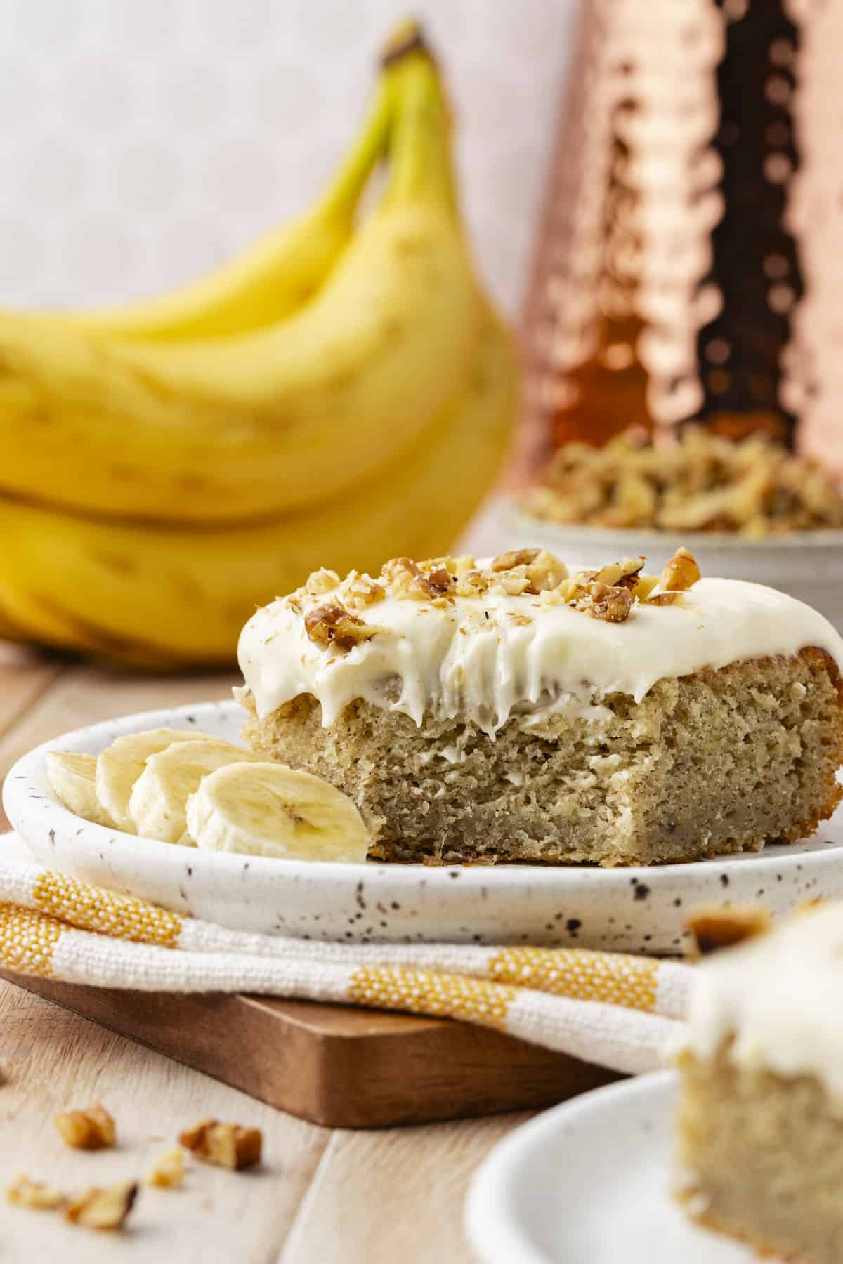 a slice of banana cake on a small plate sitting on top of a white and yellow striped towel on a wooden board, surrounded by more cake, a bowl of walnuts, and whole bananas