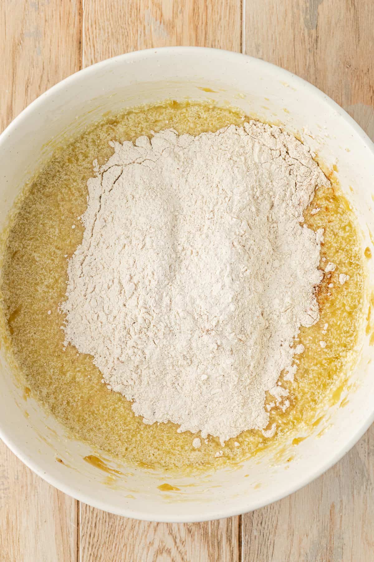 a white bowl on a wood surface with wet ingredients for banana cake and a dry flour mixture sitting on top not yet mixed in