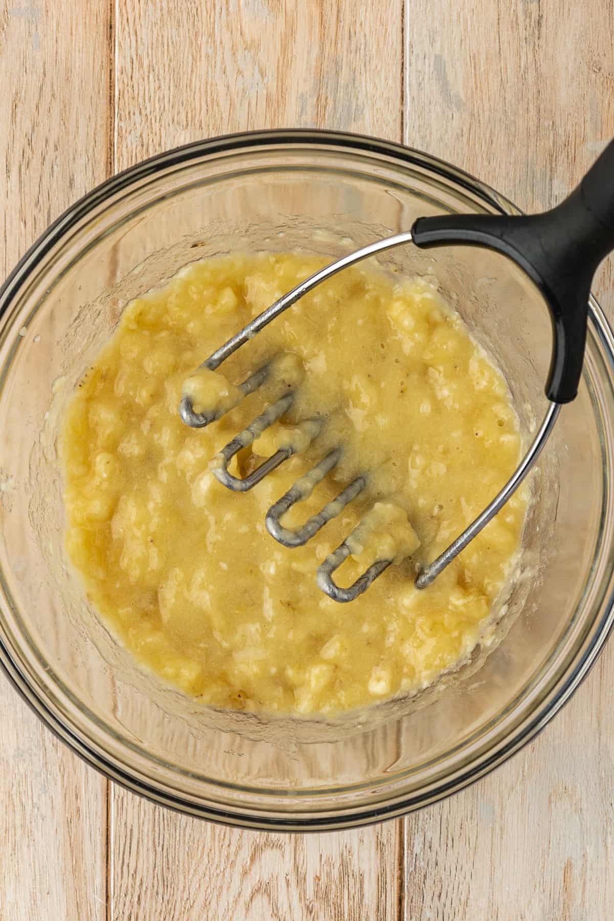 a glass bowl full of mashed banana with a masher leaning in the bowl, sitting on a wooden surface