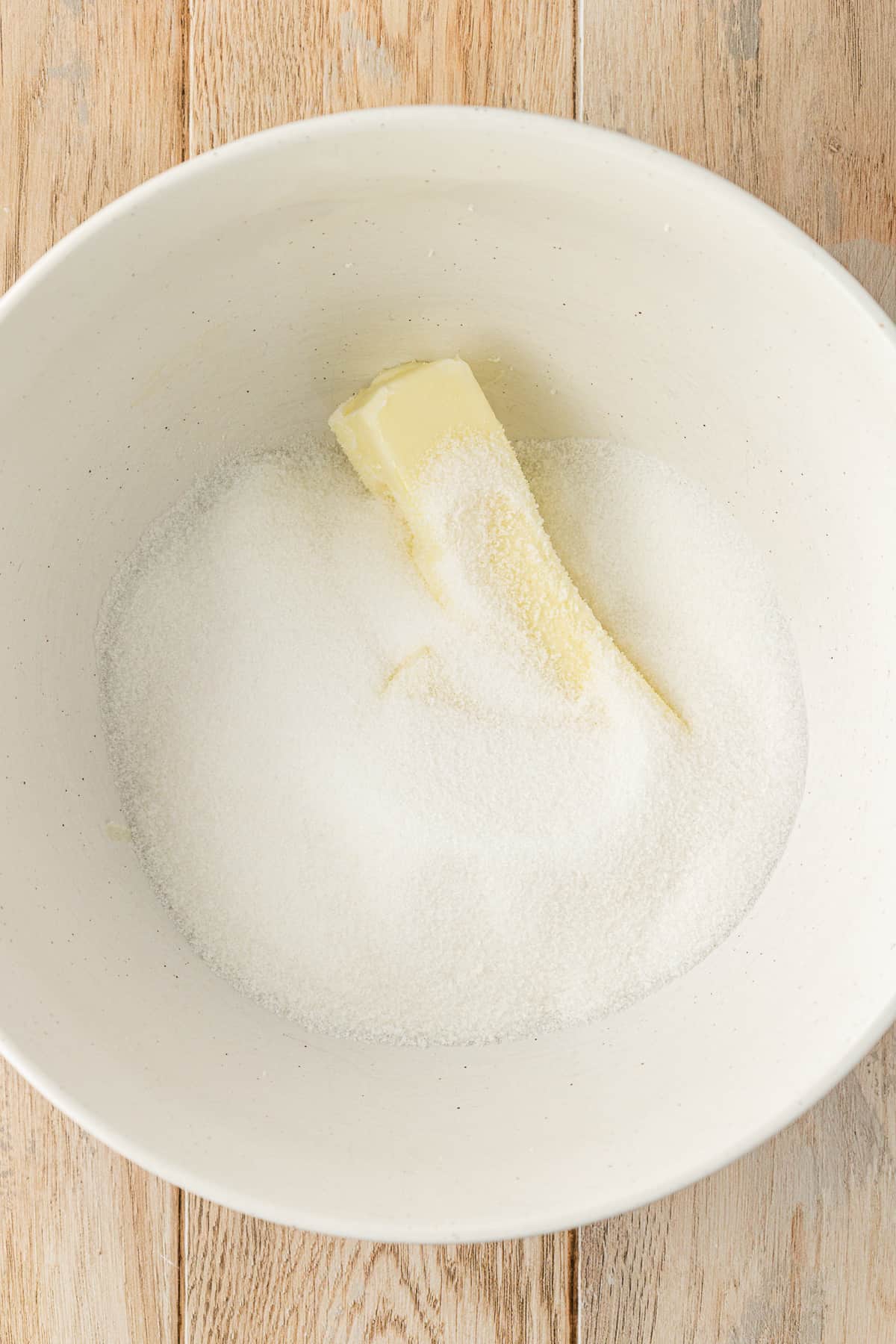 a white bowl on a wood surface with granulated sugar and a stick of butter in it