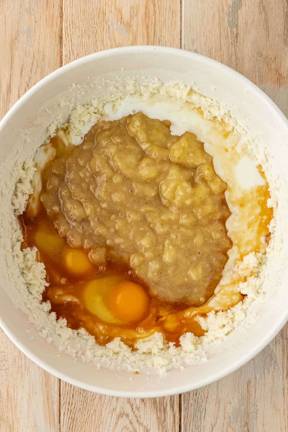 a white bowl with a butter and sugar mixture, mashed bananas, eggs, buttermilk and vanilla extract that has not been mixed together yet