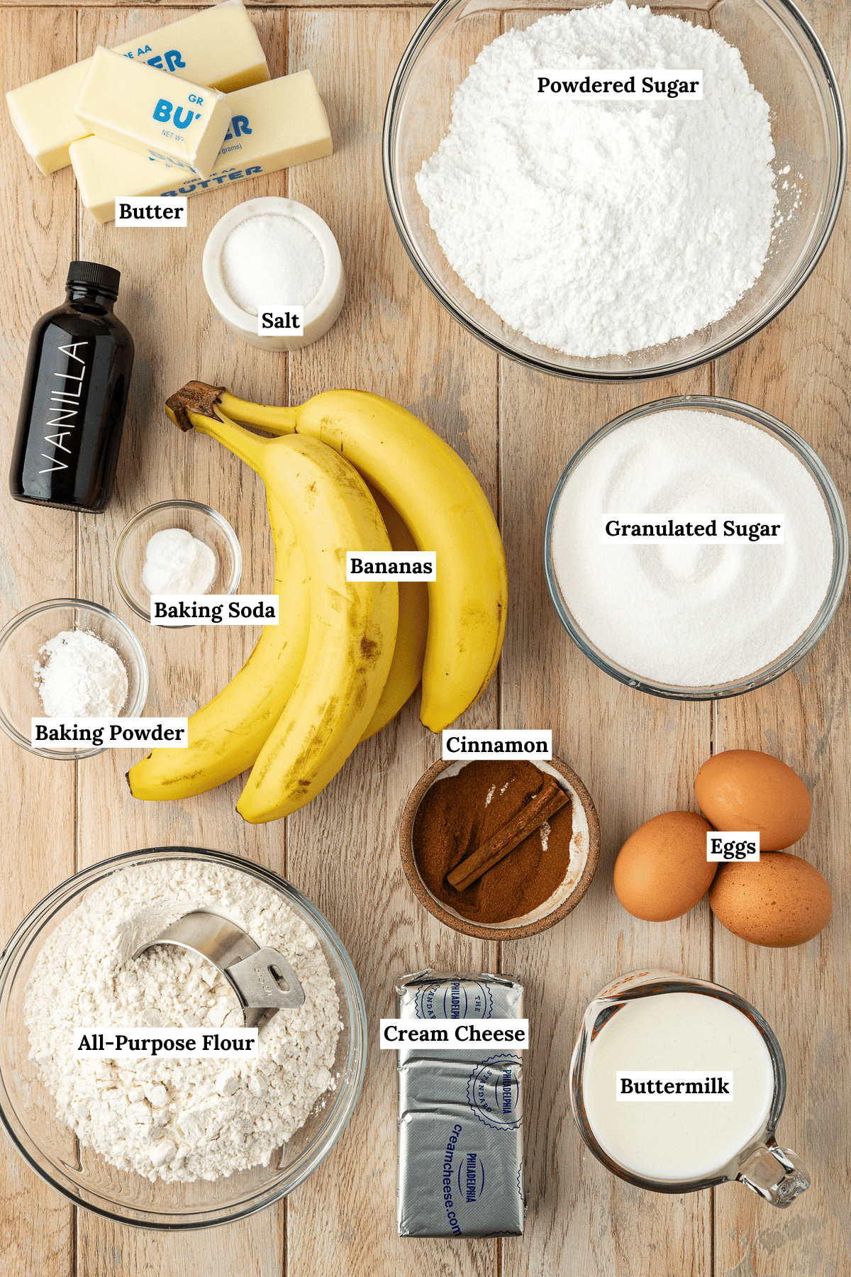 over head view of the ingredients for banana cake on a wood surface including a bowl of powdered sugar, a bowl of granulated sugar, two and a half sticks of butter, a small white dish of salt, a bottle of vanilla extract, a small bowl of baking soda, a small bowl of baking powder, a bowl of cinnamon, three large eggs, four whole bananas, a bowl of all-purpose flour, a block of cream cheese and a measuring cup of buttermilk