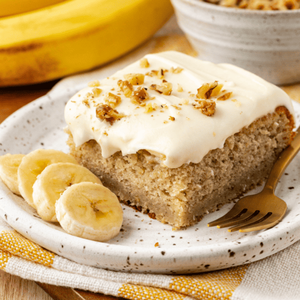 a slice of banana cake with one bite missing on a small plate with fresh banana slices, on top of a yellow and white striped kitchen towel with whole bananas and bowl of crumb topping in the background