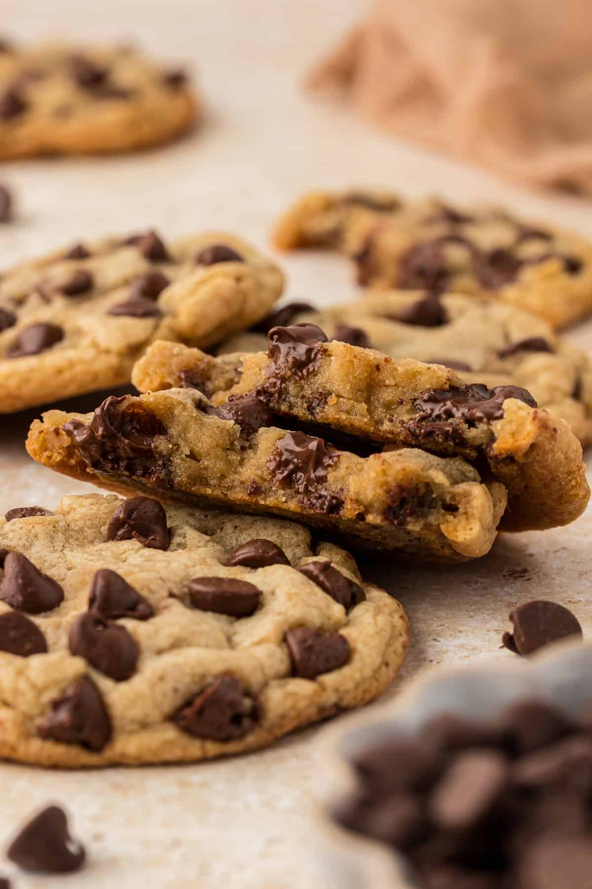 brown butter chocolate chip cookies scattered around, with one cookie broken in half and stacked leaning on another cookie exposing the inside with melted chocolate chips and more chocolate chips sprinkled around