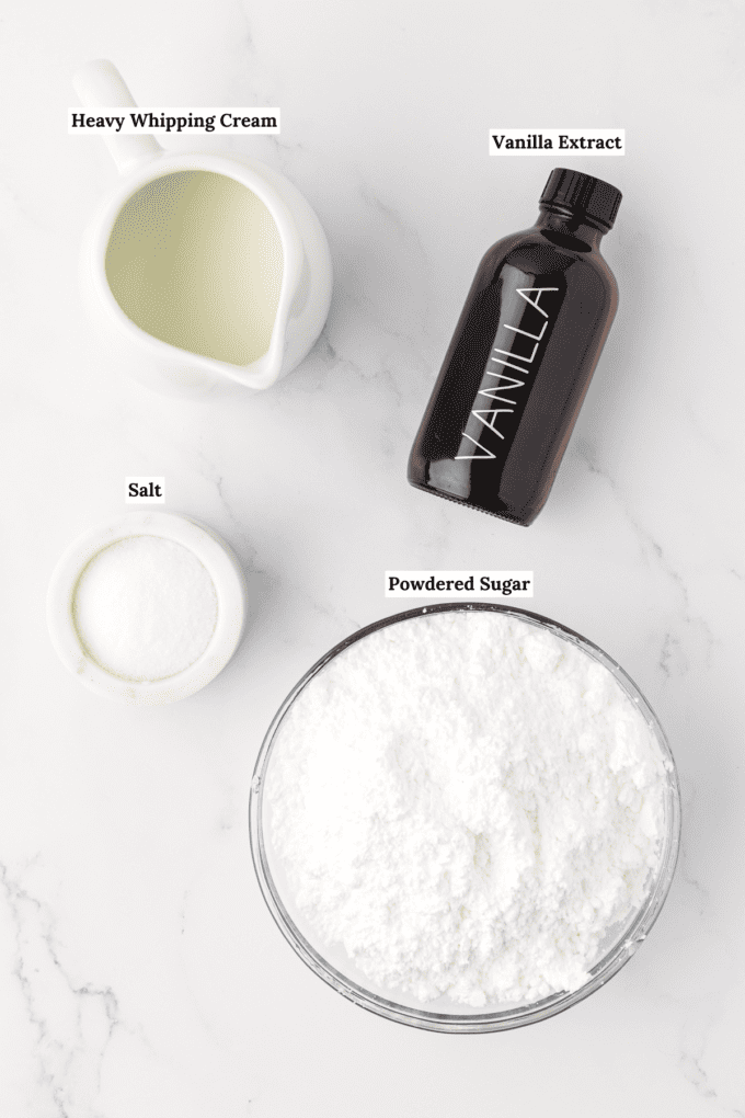 overhead view of the ingredients for vanilla glaze including a white dish full of heavy whipping cream, a bottle of vanilla extract, a small white bowl of salt and a large glass bowl of powdered sugar
