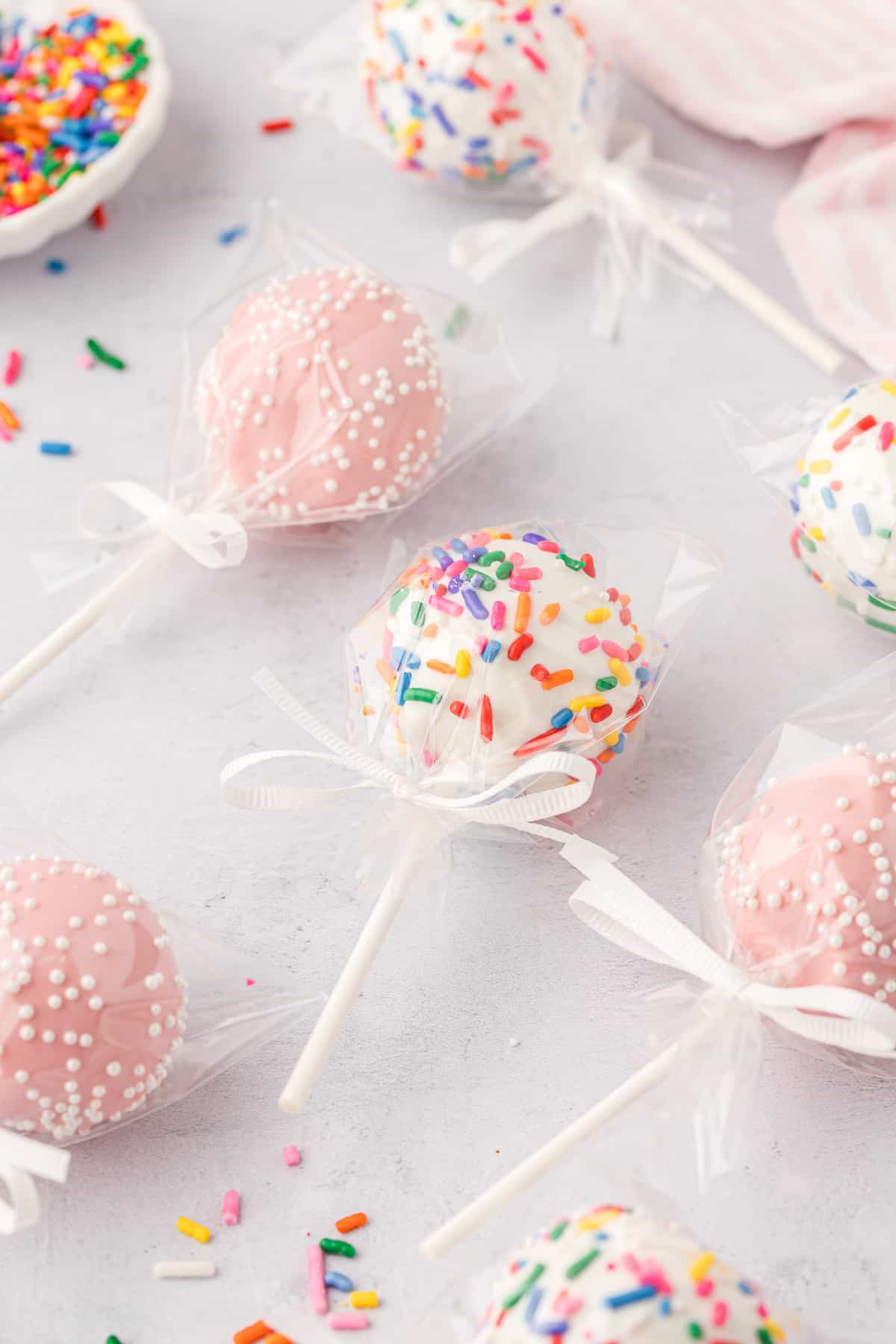 cake pops individually wrapped in plastic with white bows laying on a light grey surface, some pink with white sprinkles and some white with rainbow sprinkles, with rainbow sprinkles scattered around