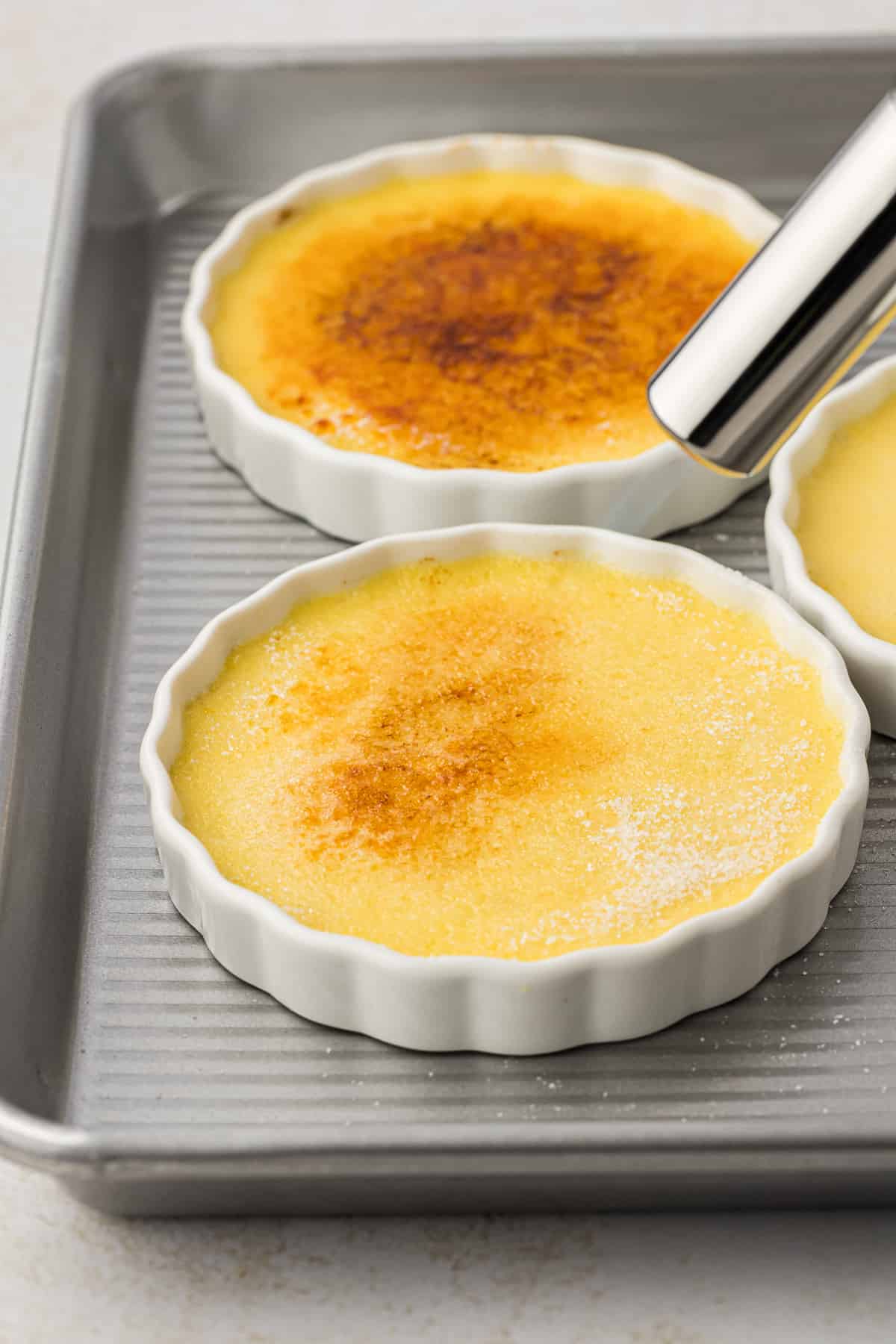 ramekins of creme brulee on a baking sheet being caramelized on top with a blow torch