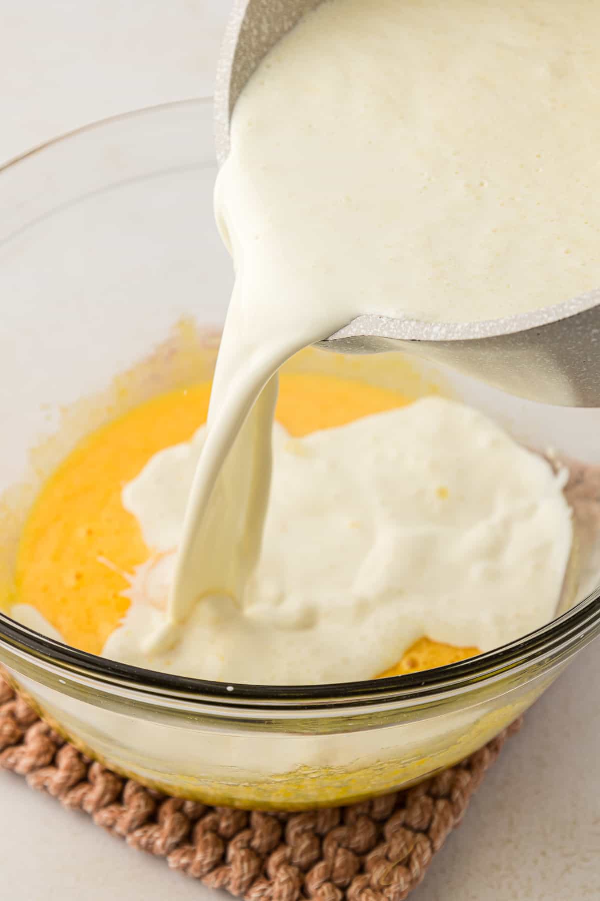 a cream mixture being poured from a pot into a clear glass bowl with a egg and sugar mixture in it