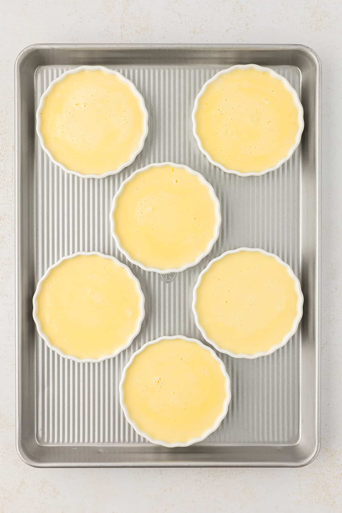 over head view of a baking sheet with six white ramekins on it full of a cream mixture for creme brulee
