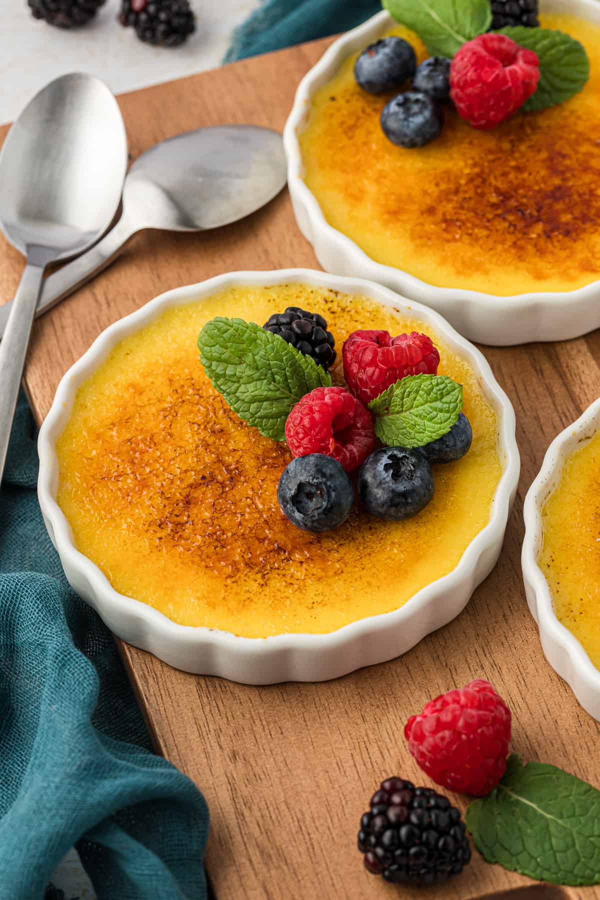 creme brulee in white ramekins on a wooden cutting board with two spoons, on top of a dark teal towel with fresh berries and mint leaves topping the creme brulee and more sprinkled around