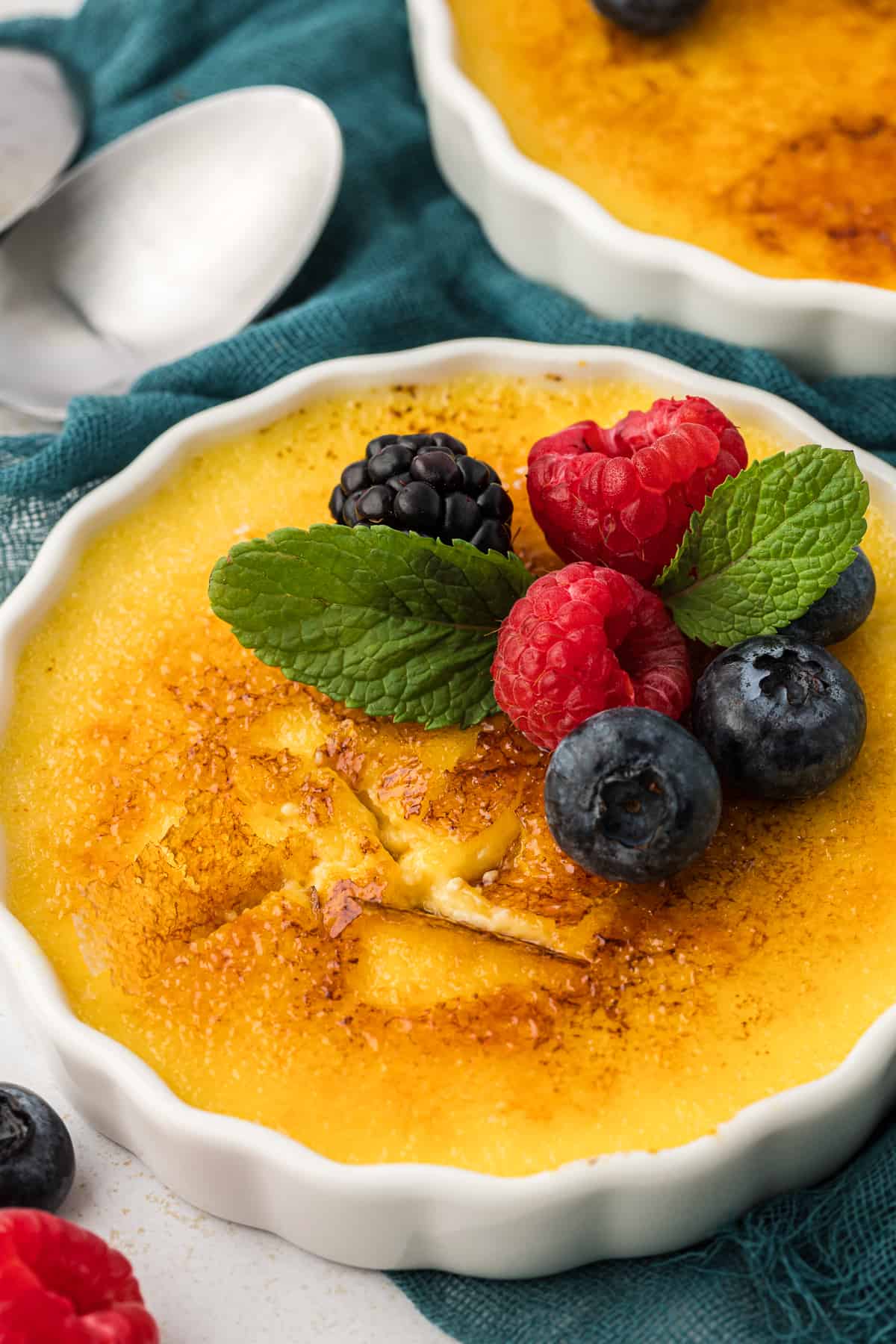 a white ramekin of creme brulee topped with fresh berries and mint leaves on top of a dark teal towel surrounded by more fresh berries, spoons and another dish of creme brulee in the background