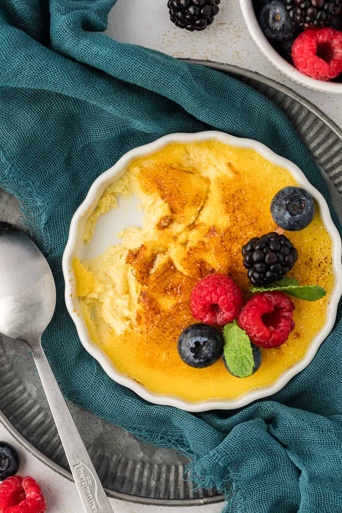 a white ramekin of creme brulee topped with fresh berries and mint leaves with some bites missing, sitting on a dark teal towel on a dark grey plate with a spoon with more fresh berries around the plate