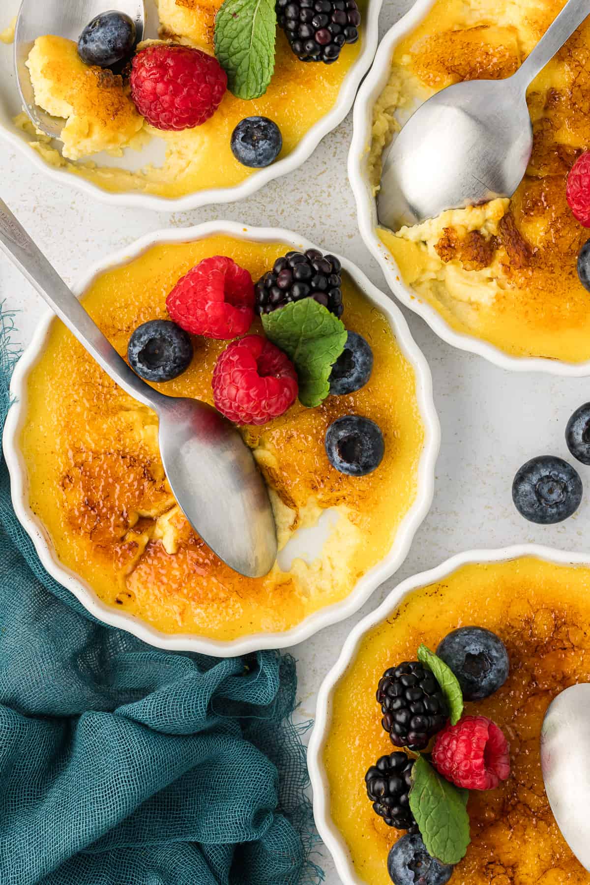 four white ramekins of creme brulee with spoons in them, some with bites missing out, topping with fresh berries and mint leaves beside a dark teal towel and more fresh berries scattered around