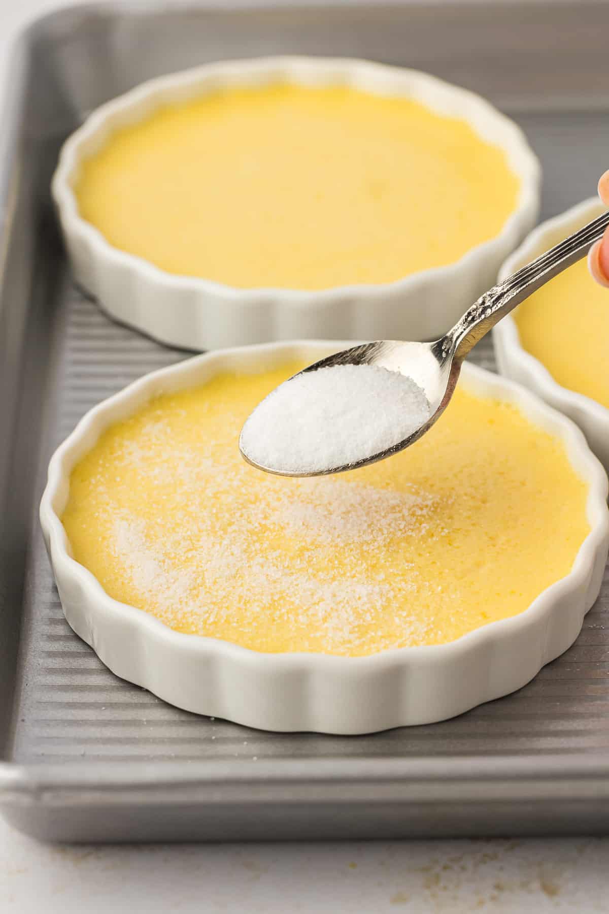 ramekins of creme brulee on a baking sheet with one having a thin layer of sugar added to the top with a spoon