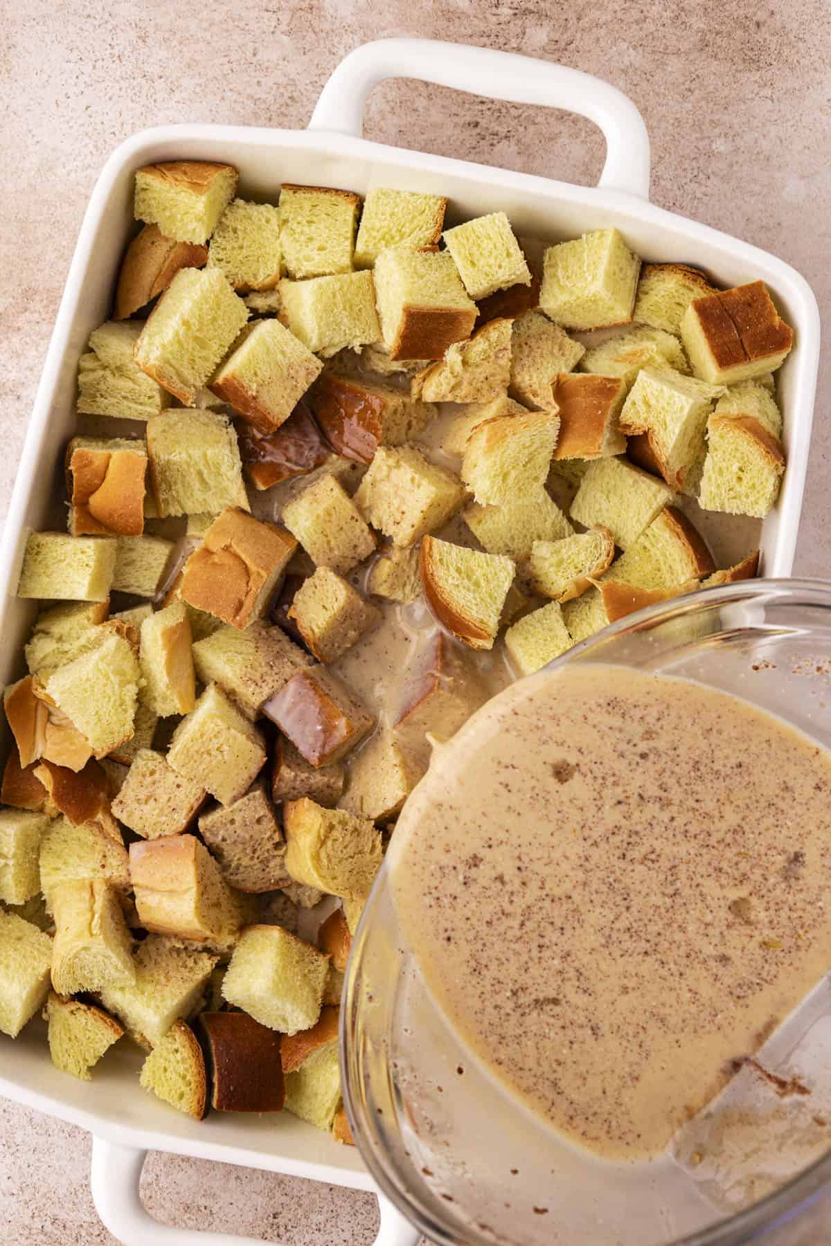 a white baking dish full of bread cubes with a liquid mixture being poured over top of it