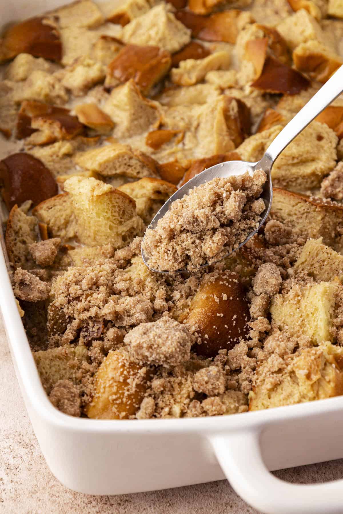 a white baking dish with french toast casserole mixture in it being sprinkled over the top with a spoon of crumb topping