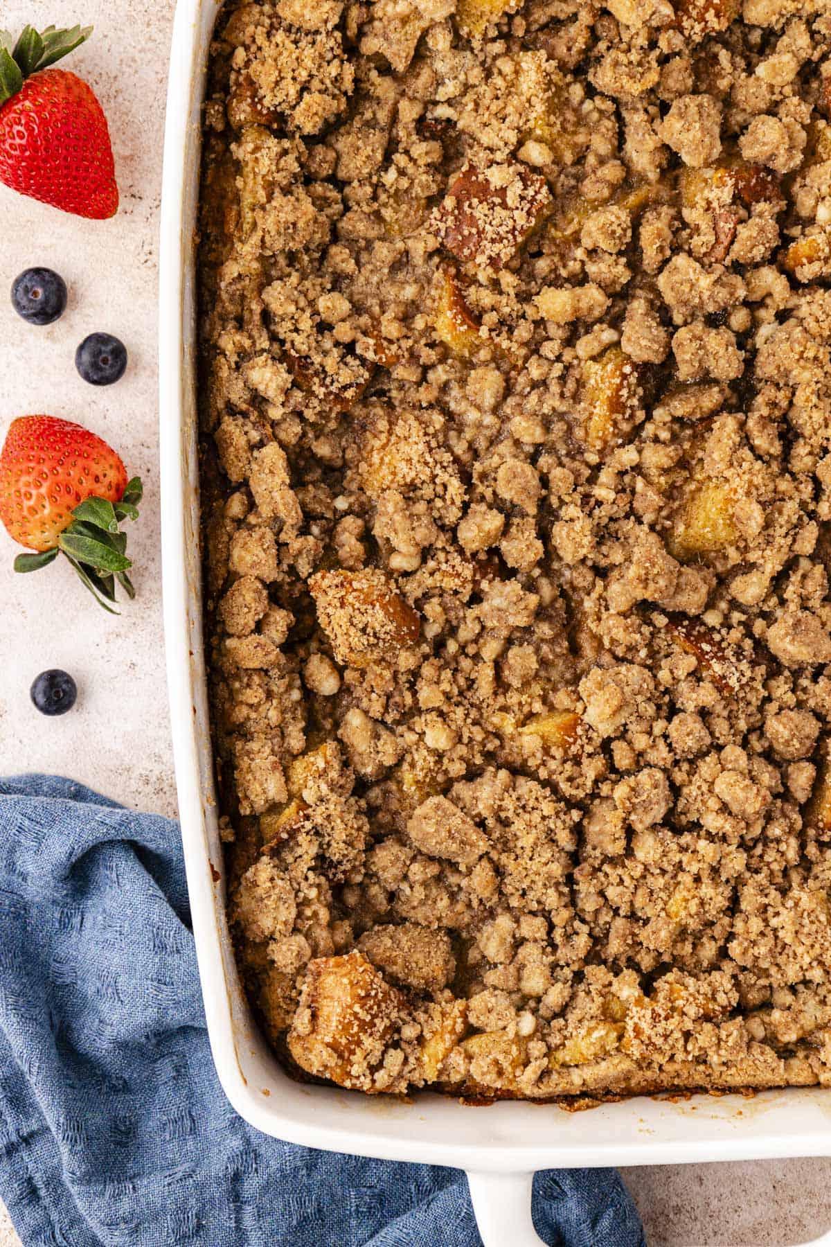 a large white baking dish of french toast casserole with crumb topping beside a blue kitchen towel and fresh blueberries and strawberries scattered around
