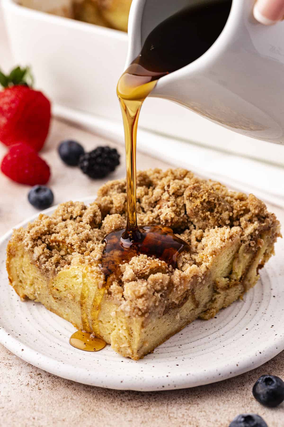 a slice of french toast casserole on a small plate with maple syrup being poured over the top and fresh berries scattered around